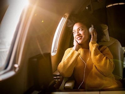 Eine junge Frau schaut mit Kopfhörern aus dem Fenster eines Flugzeugs, wobei das Sonnenlicht durch das Fenster fällt.